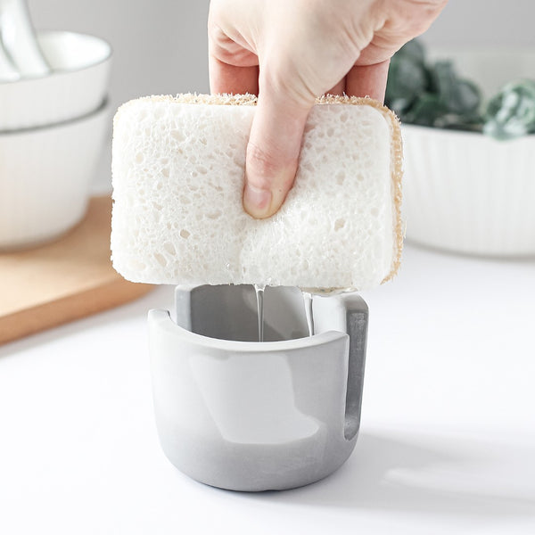 Hand using a white sponge to clean a gray kitchen tool on a light surface.