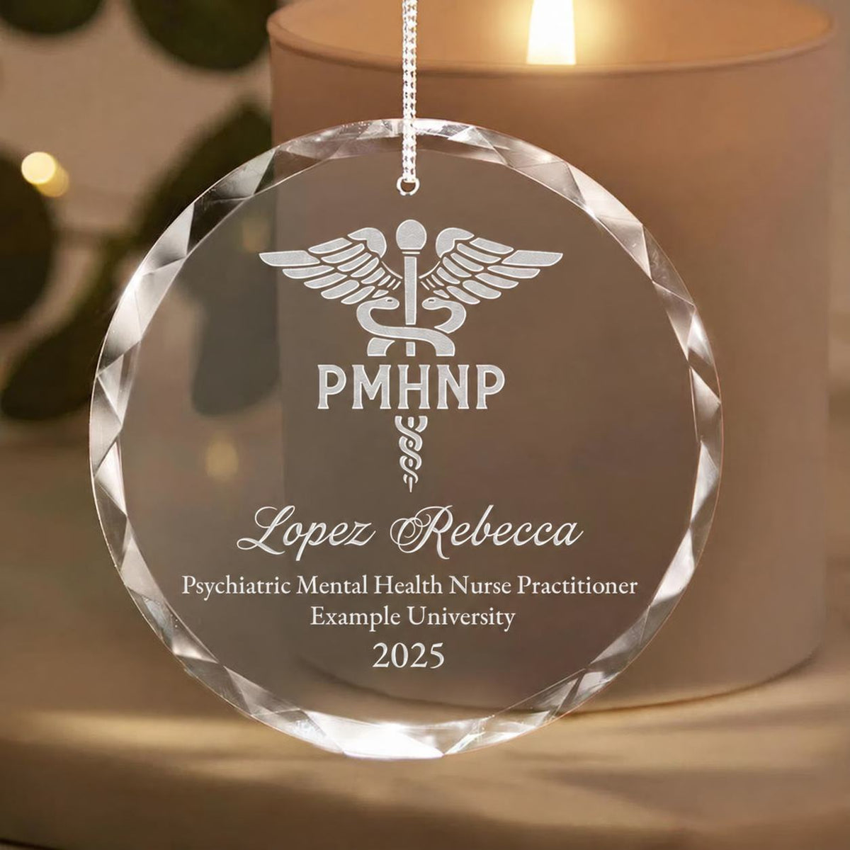 A round clear hanging ornament with a faceted crystal edge, engraved with a medical caduceus symbol and the text. It hangs in front of a softly blurred beige candle holder with warm light.
