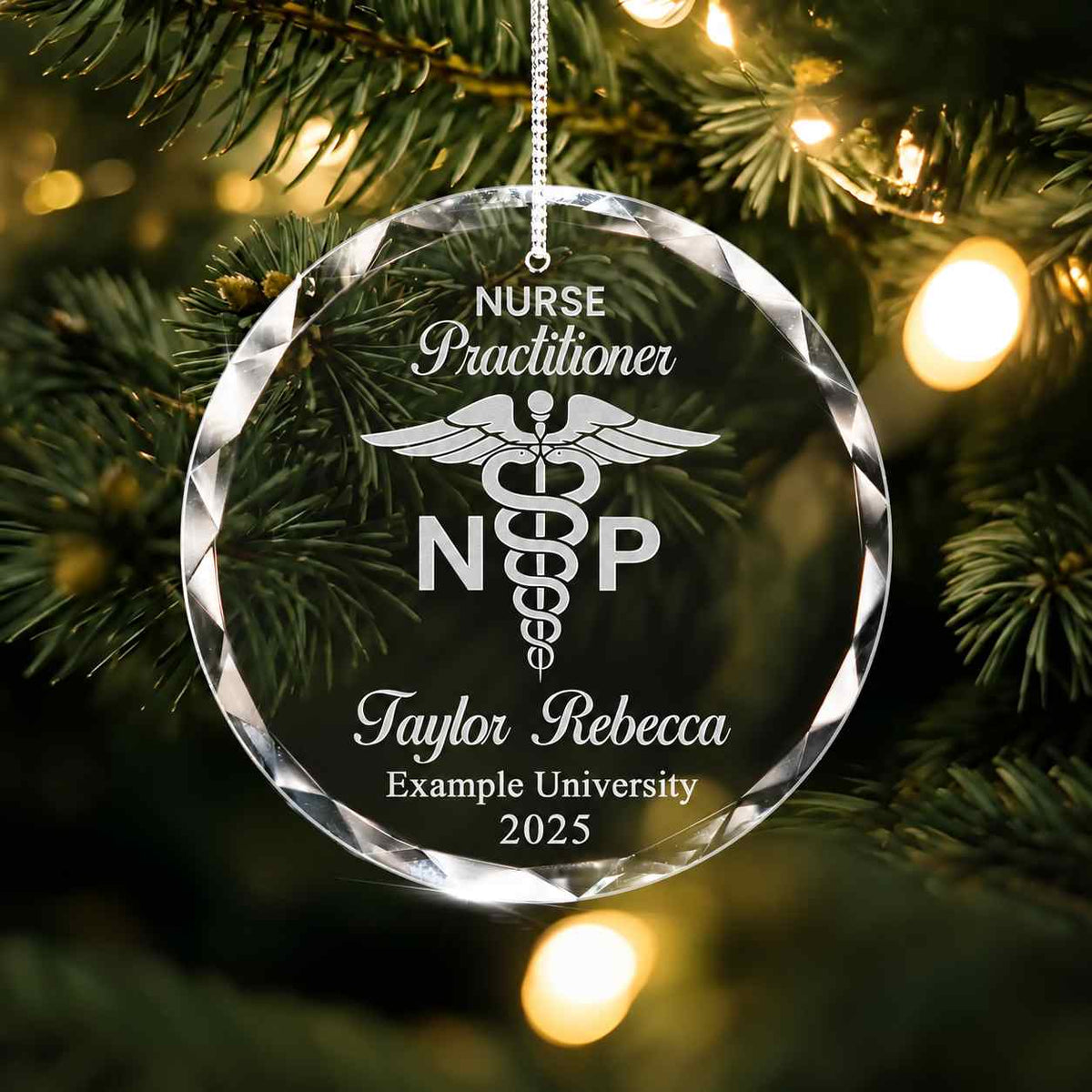 A personalized clear glass or acrylic Christmas ornament shaped like a faceted round disc, hanging from a string in a Christmas tree. The ornament is engraved with the words 'Nurse Practitioner', a caduceus symbol, 'NRP', the name 'Taylor Rebecca', 'Example University', and '2025'.