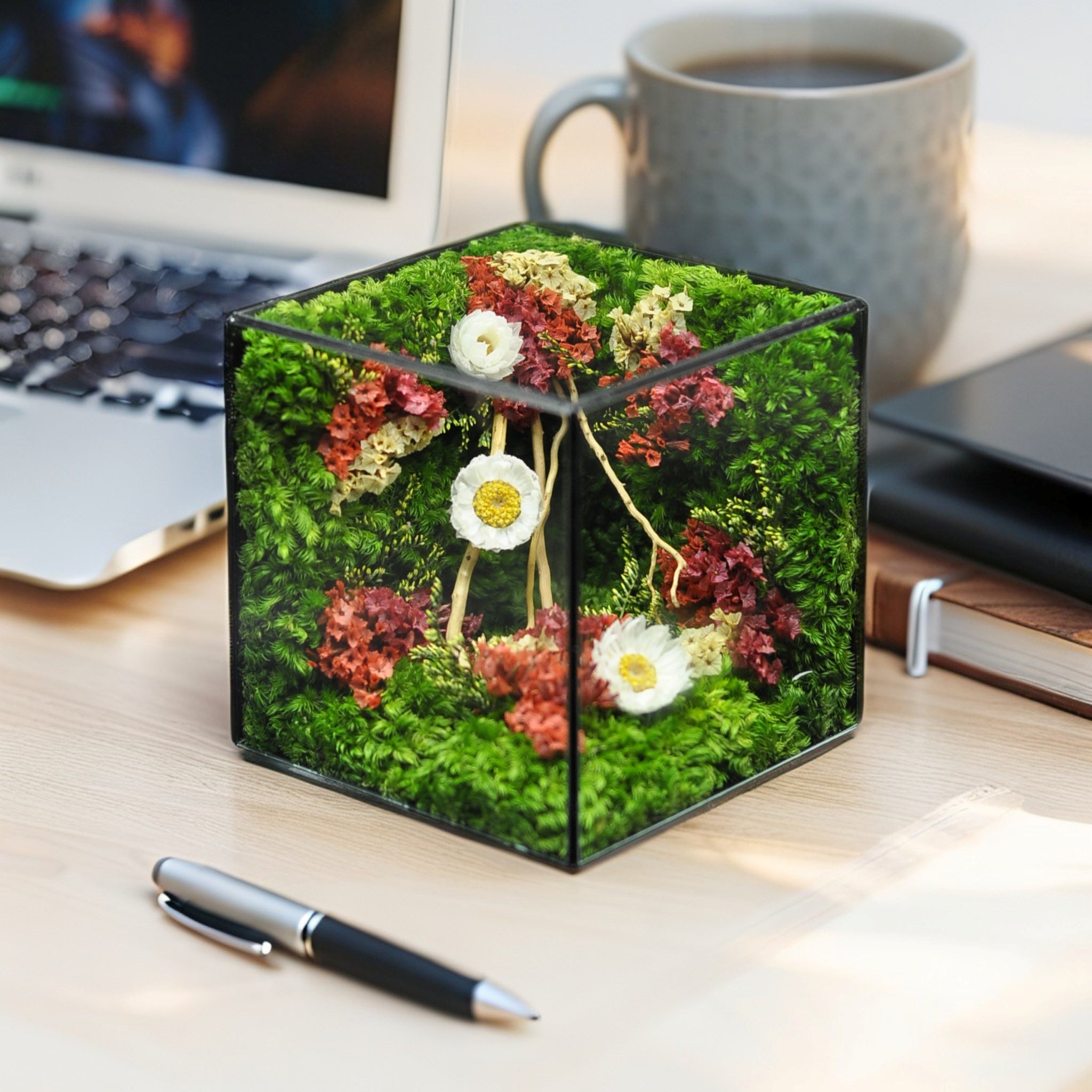 Decorative moss cube with flowers on a desk next to a laptop, mug, and pen.