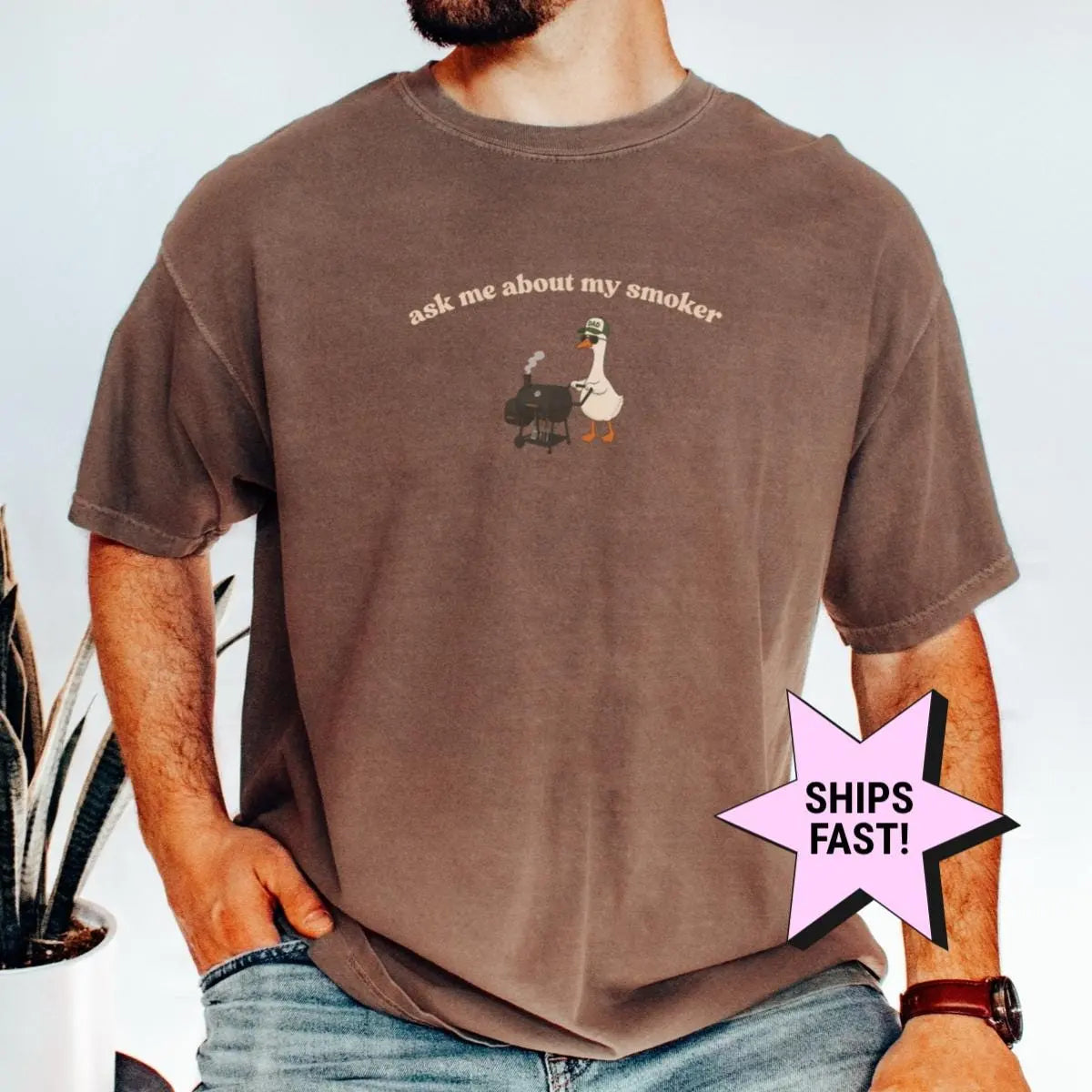 Man in an Espresso Comfort Colors tee, hand in pocket, with a goose at a smoker grill under ask me about my smoker
