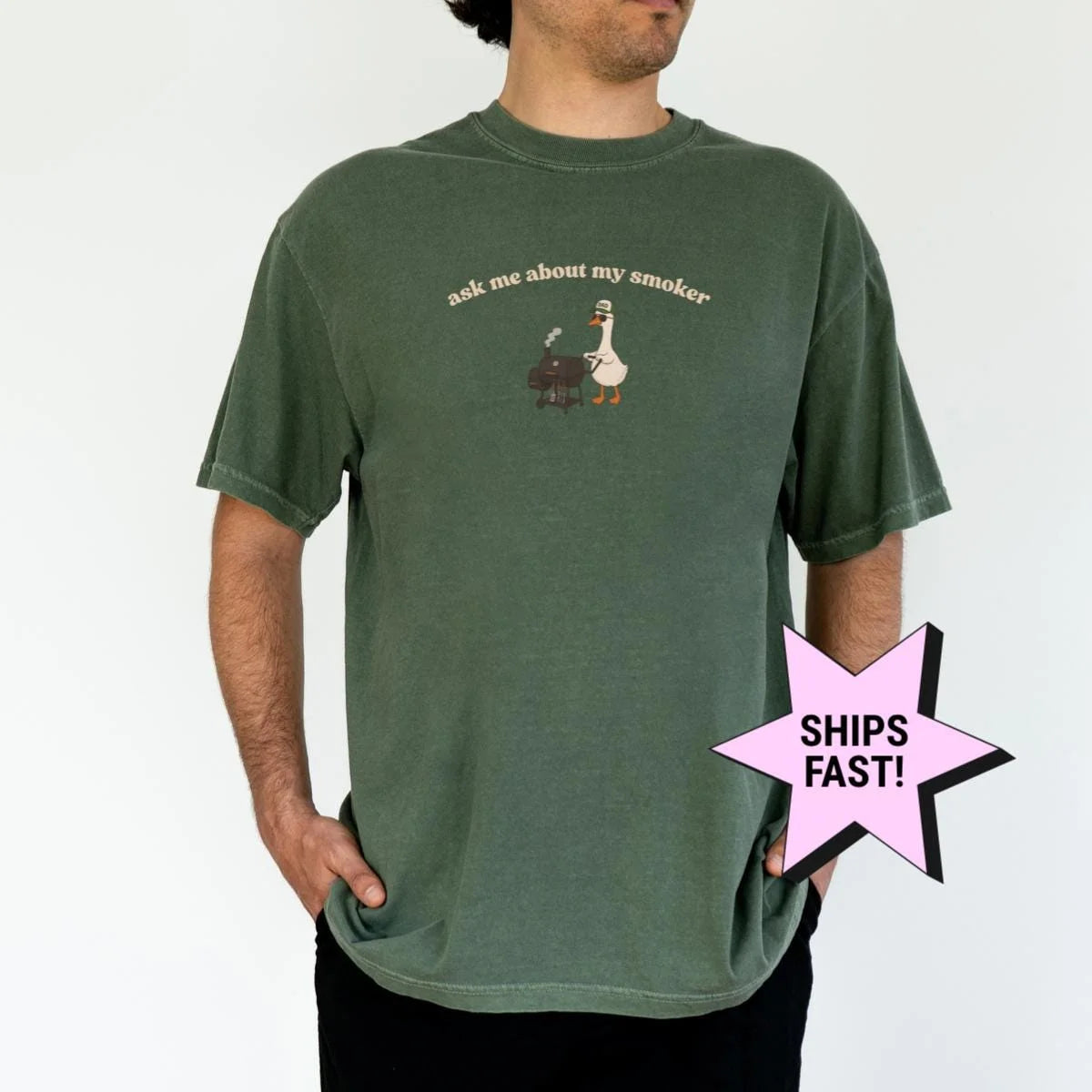 Man in a Moss green Comfort Colors tee with a small goose at a smoker grill under ask me about my smoker, in tan