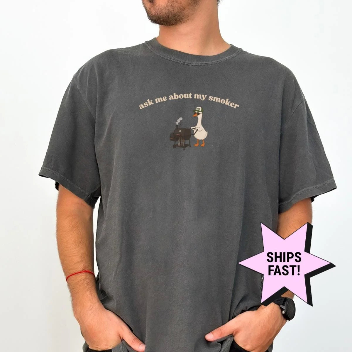 Man in a Pepper Comfort Colors tee with a small goose at a smoker grill under arched ask me about my smoker text