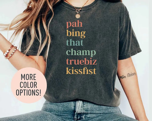 Woman in a dark gray Comfort Colors tee and jeans, bracelets on her wrist, with six stacked ASL slang words in coral and sage