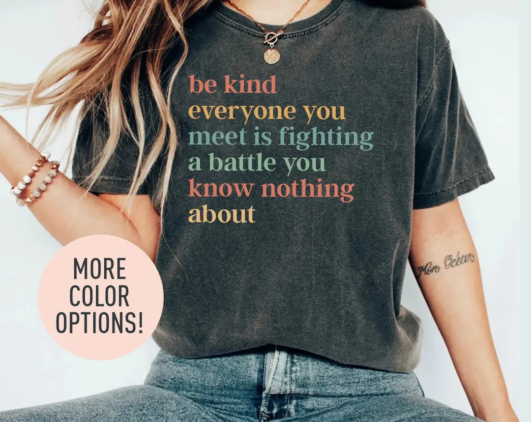 Woman in a dark gray Comfort Colors tee and jeans, bracelets on her wrist, with a six-line be kind quote in coral and sage