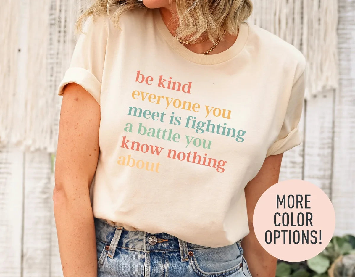 Woman in a natural cream tee with rolled sleeves and jeans, the be kind battle quote in coral, mustard and sage serif