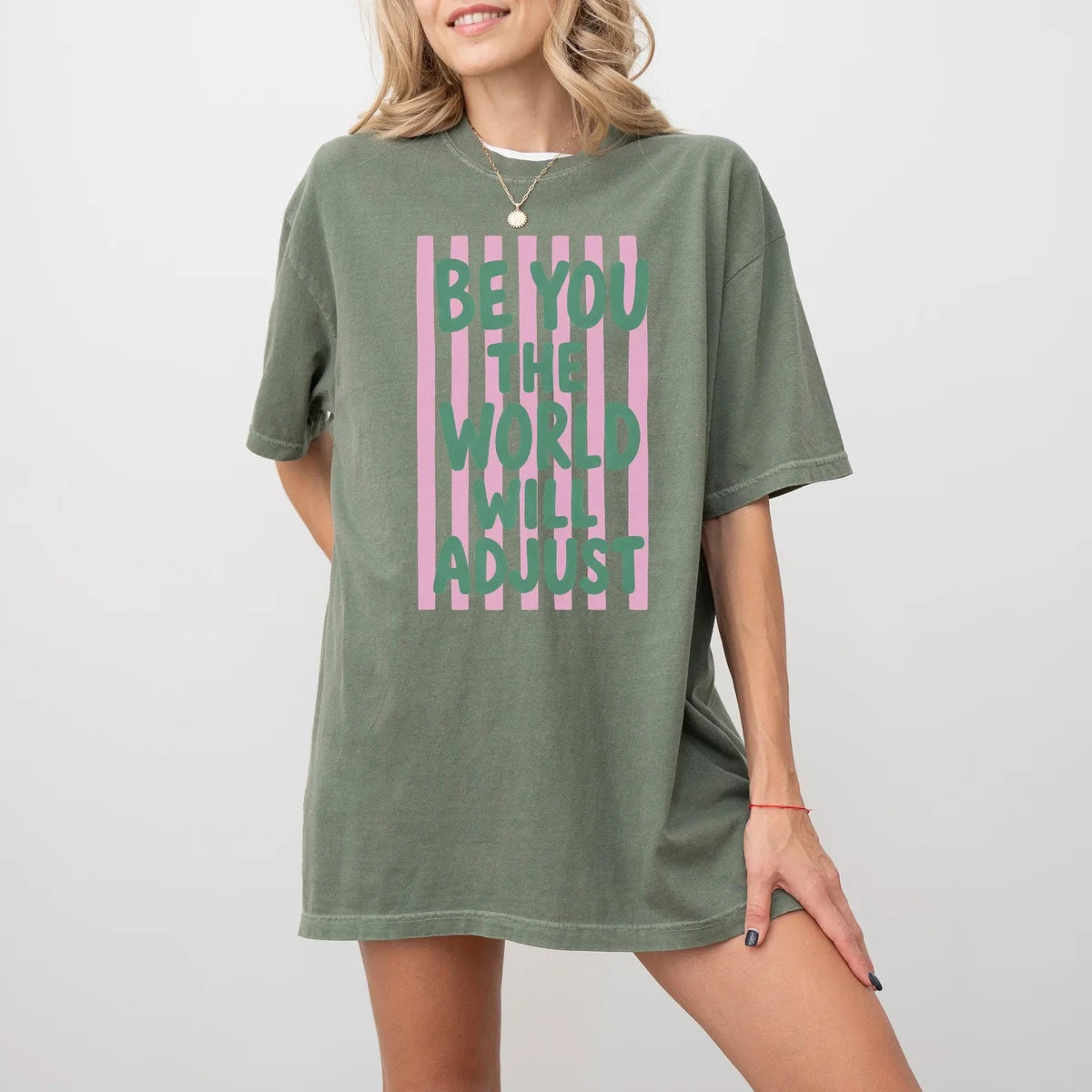 Woman in a Moss green Comfort Colors tee, hand on hip, with green be you the world will adjust text over pink stripes