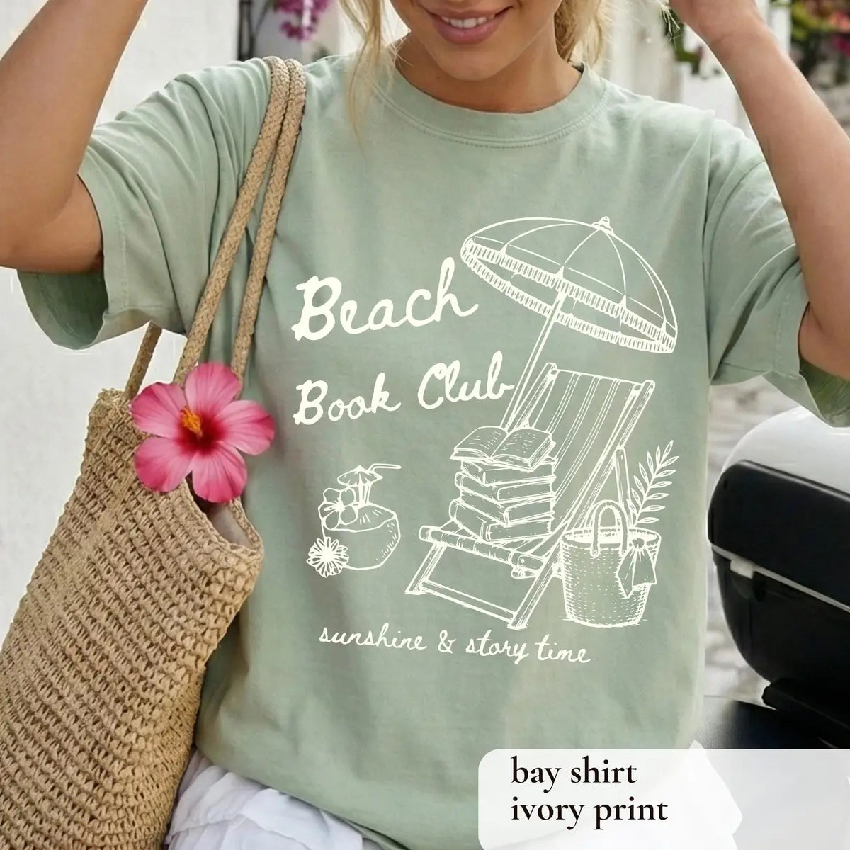 Woman in a Bay sage Comfort Colors tee with a straw tote and hibiscus, ivory beach book club and book-stacked chair