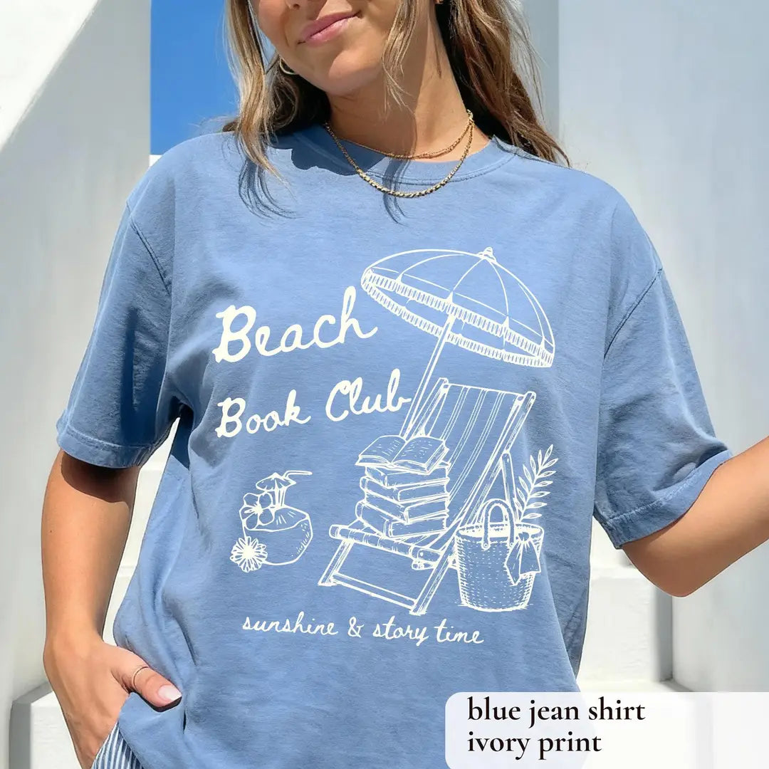 Woman in a Blue Jean Comfort Colors tee, white beach book club over a striped lounge chair with books and a tote