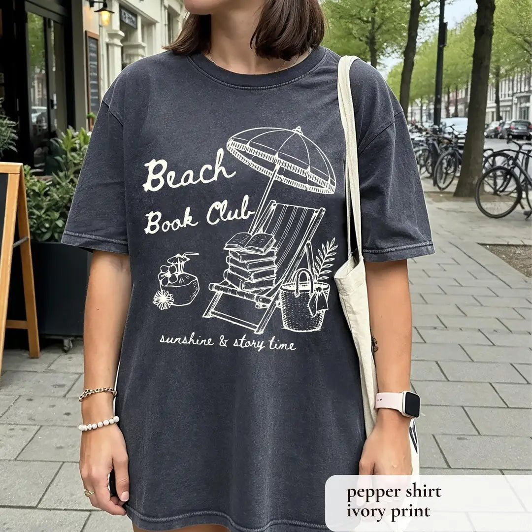 Woman in a Pepper Comfort Colors tee on a city street with a tote bag, ivory beach book club over a book-stacked chair