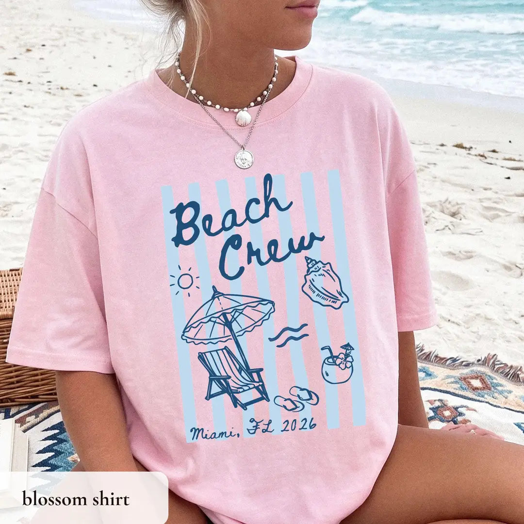 Woman in a Blossom pink Comfort Colors tee on a beach blanket, navy beach crew lettering on blue and pink stripes