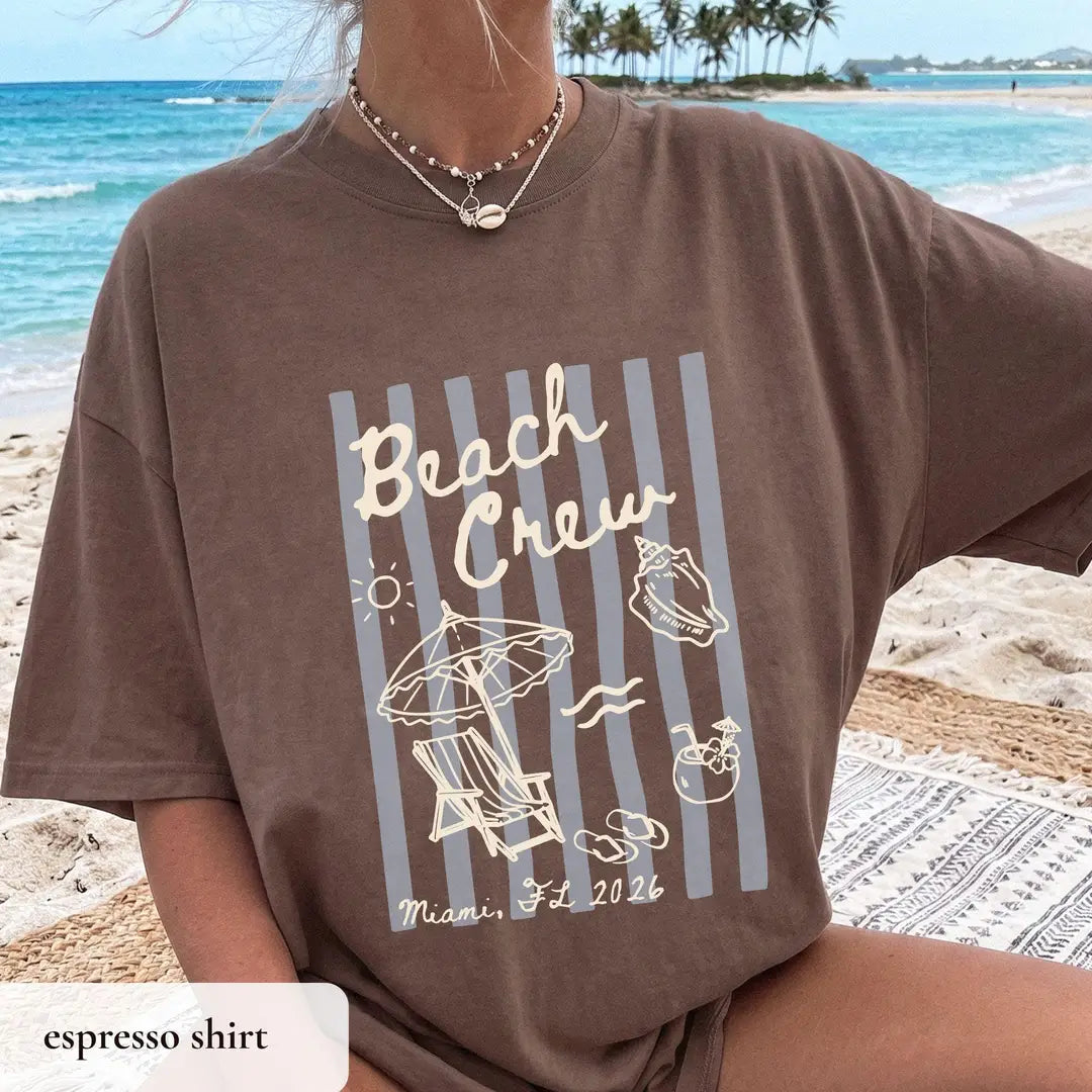 Woman in an Espresso brown Comfort Colors tee at the beach, ivory beach crew over gray-blue stripes, shell and sun