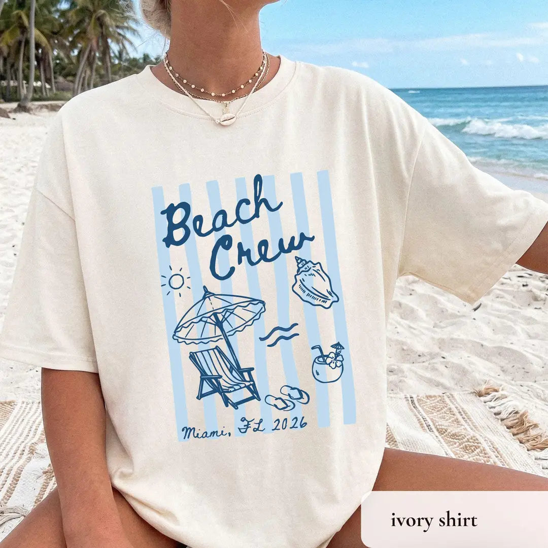 Woman in an Ivory Comfort Colors tee at the shore, blue beach crew lettering on light blue stripes, shell and umbrella