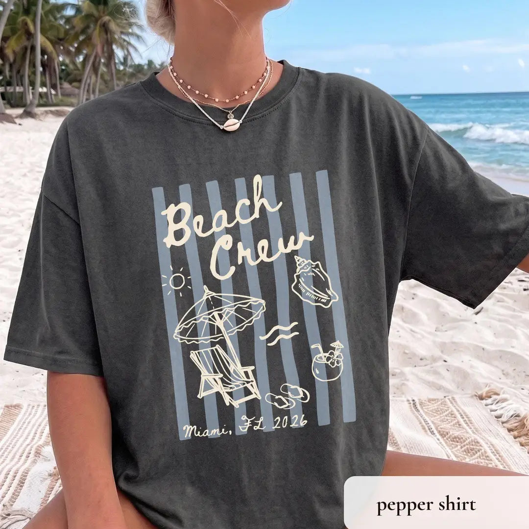 Woman in a Pepper Comfort Colors tee on a beach, cream beach crew script over blue stripes with umbrella and lounge chair