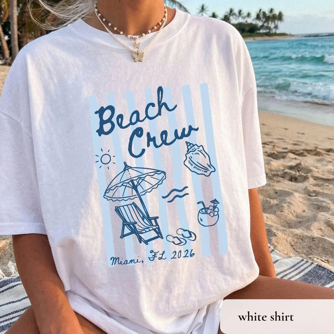 Woman in a White Comfort Colors tee by the surf, navy beach crew over striped panels, umbrella and a coconut drink