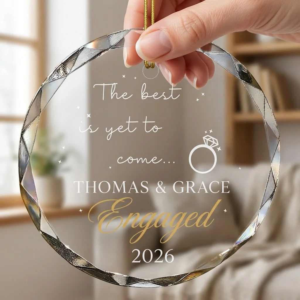 Hand holding a transparent faceted glass ornament with gold string loop showing printed diamond engagement ring line art design and crisp white lettering in cozy indoor scene