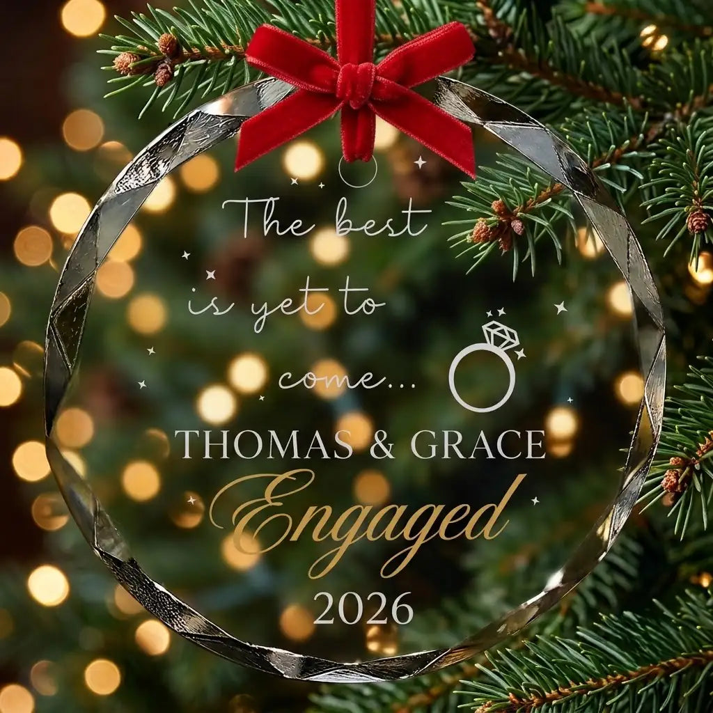 Personalized clear glass engagement ornament displayed hanging on Christmas tree branch with warm sparkling holiday lights background