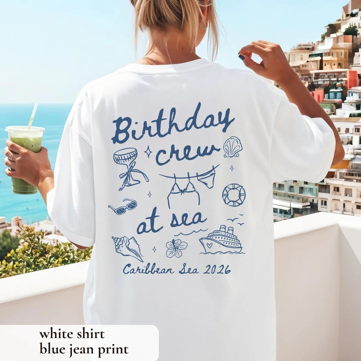 Woman from behind on a balcony above a seaside town in a White tee with Birthday crew at sea in blue and Caribbean Sea 2026