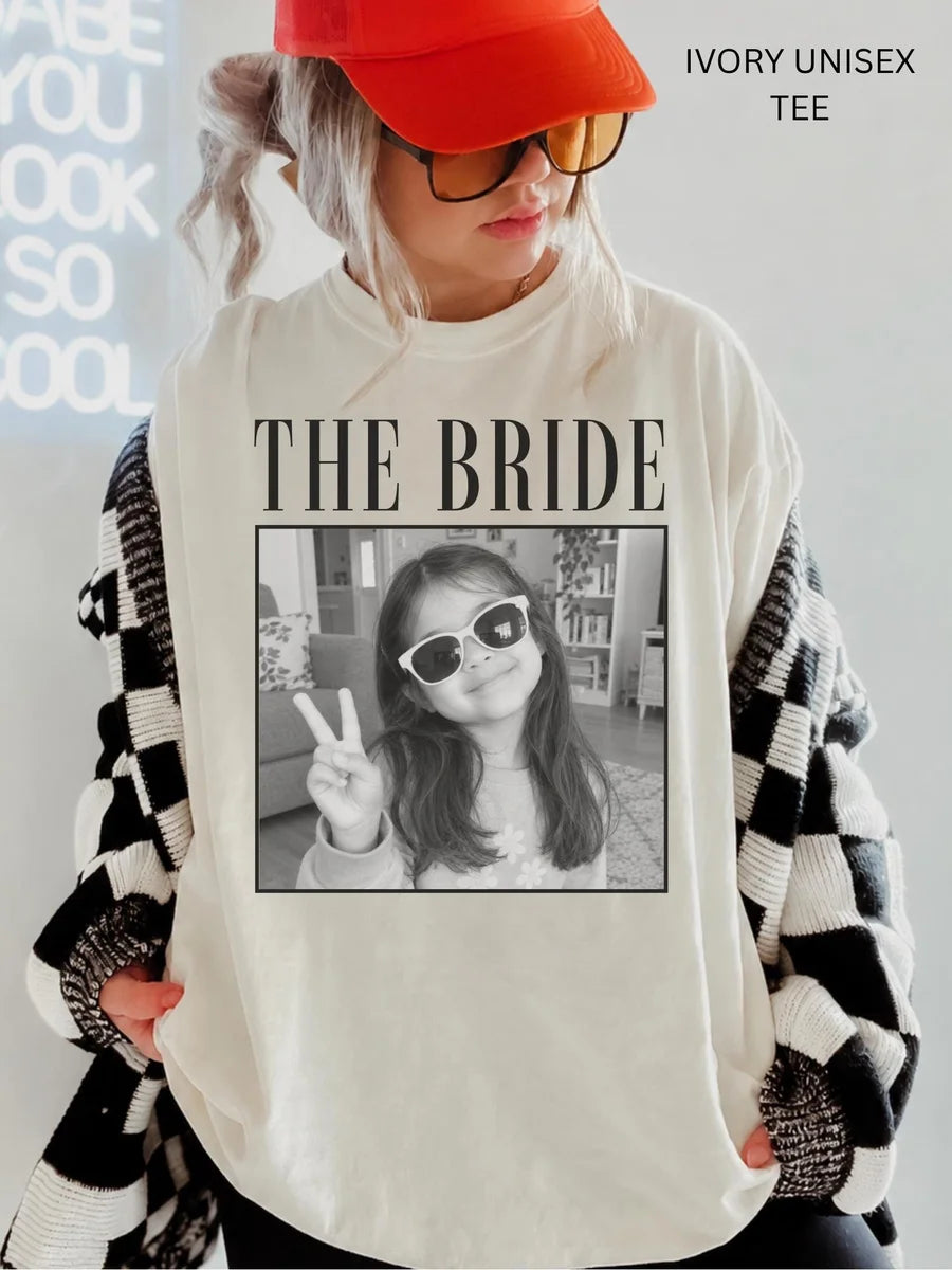 Woman in an Ivory Comfort Colors tee, red cap and checkered cardigan, the bride above a framed black and white photo