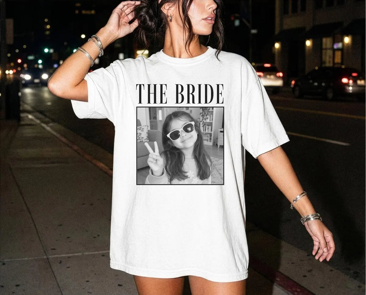Woman in a White Comfort Colors tee on a night street, bracelets, the bride in serif capitals above a framed portrait