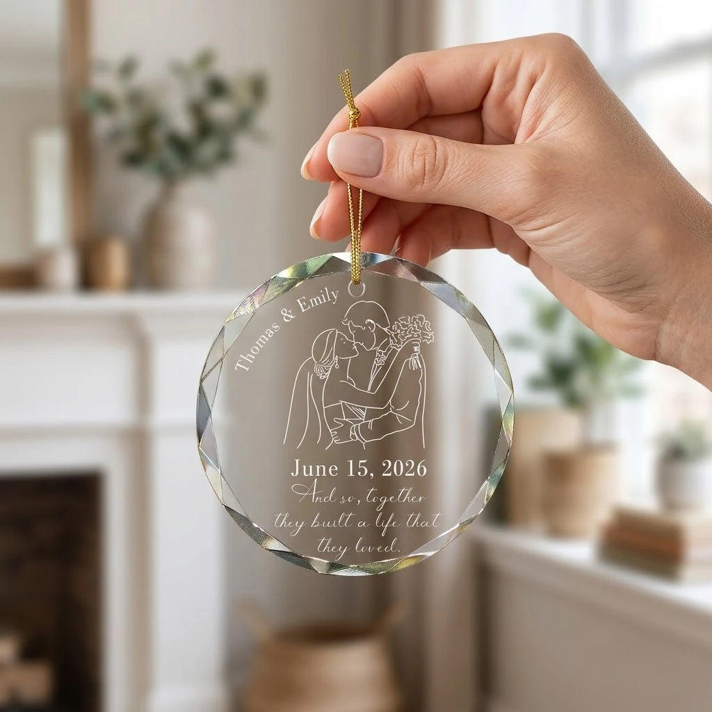 Hand holding a transparent faceted glass ornament with gold string loop showing printed wedding bride and groom line art drawing design and crisp white lettering in cozy indoor scene