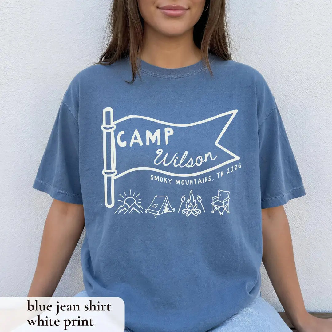 Woman in a Blue Jean Comfort Colors tee seated against a white wall, white Camp Wilson pennant with tent, campfire and chair