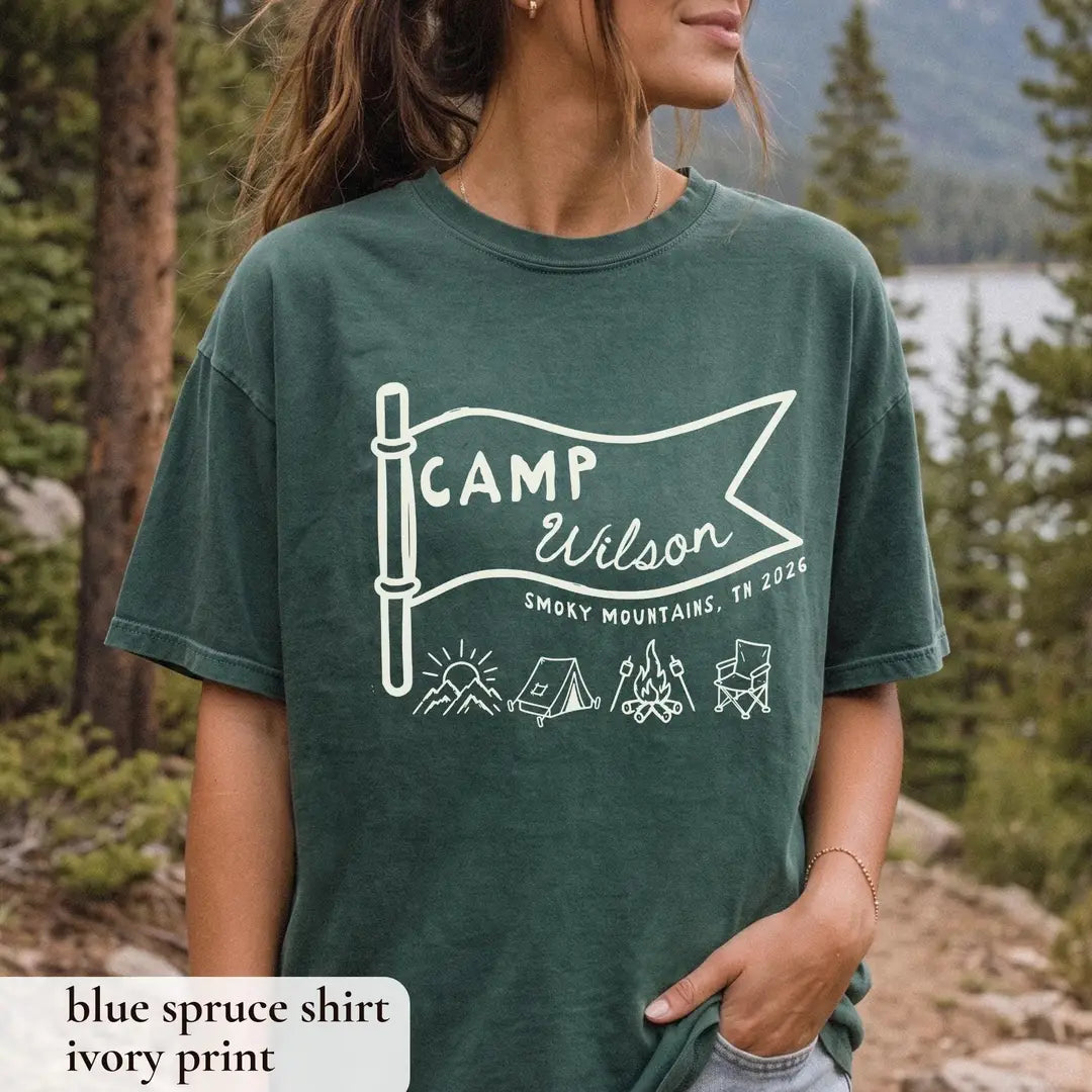 Woman in a Blue Spruce Comfort Colors tee by a mountain lake and pines, ivory Camp Wilson pennant flag with camping icons