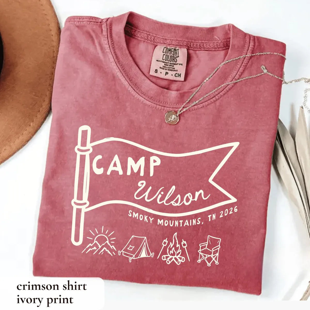 Folded Crimson Comfort Colors tee with an ivory Camp Wilson pennant flag, sunrise, tent, campfire and chair, tan hat nearby