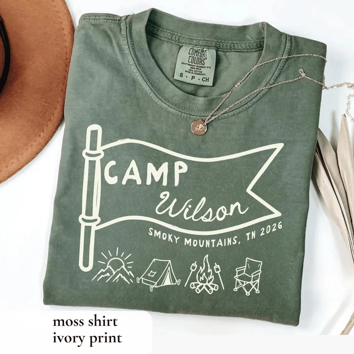 Folded Moss green Comfort Colors tee with an ivory Camp Wilson pennant flag, sun, tent, campfire and chair, necklace