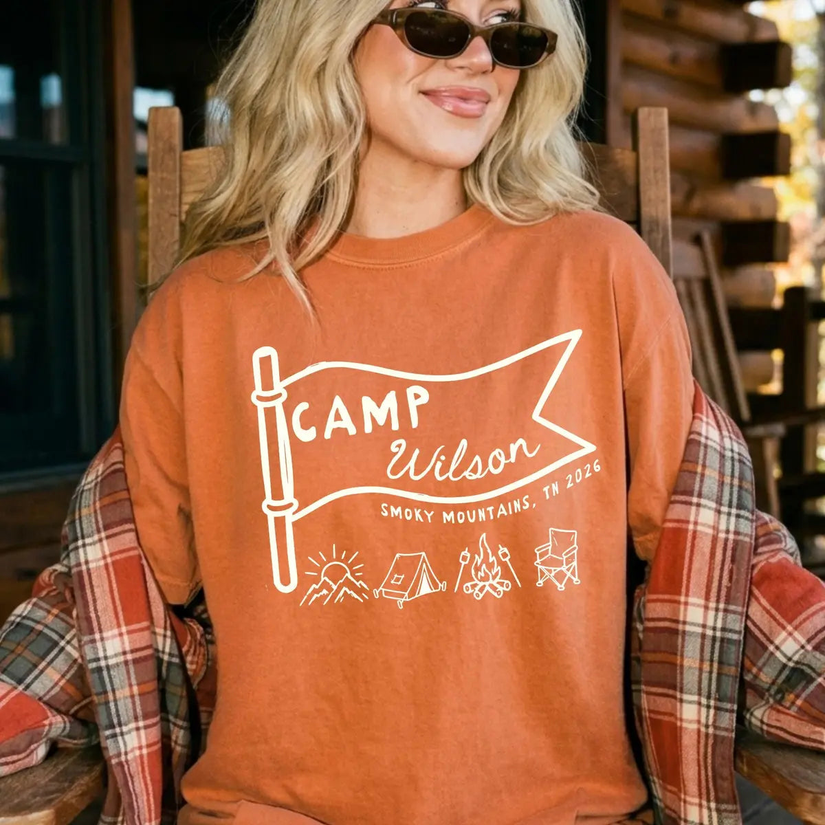 Smiling woman in sunglasses and a Yam orange Comfort Colors tee on a cabin porch, ivory Camp Wilson pennant flag with tent