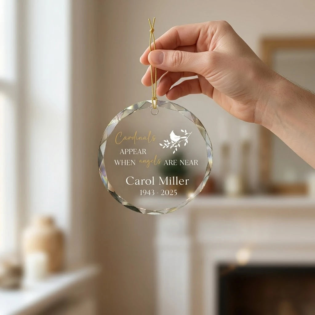 Hand holding a transparent faceted glass ornament with gold string loop showing printed cardinal bird on branch design and crisp white lettering in cozy indoor scene