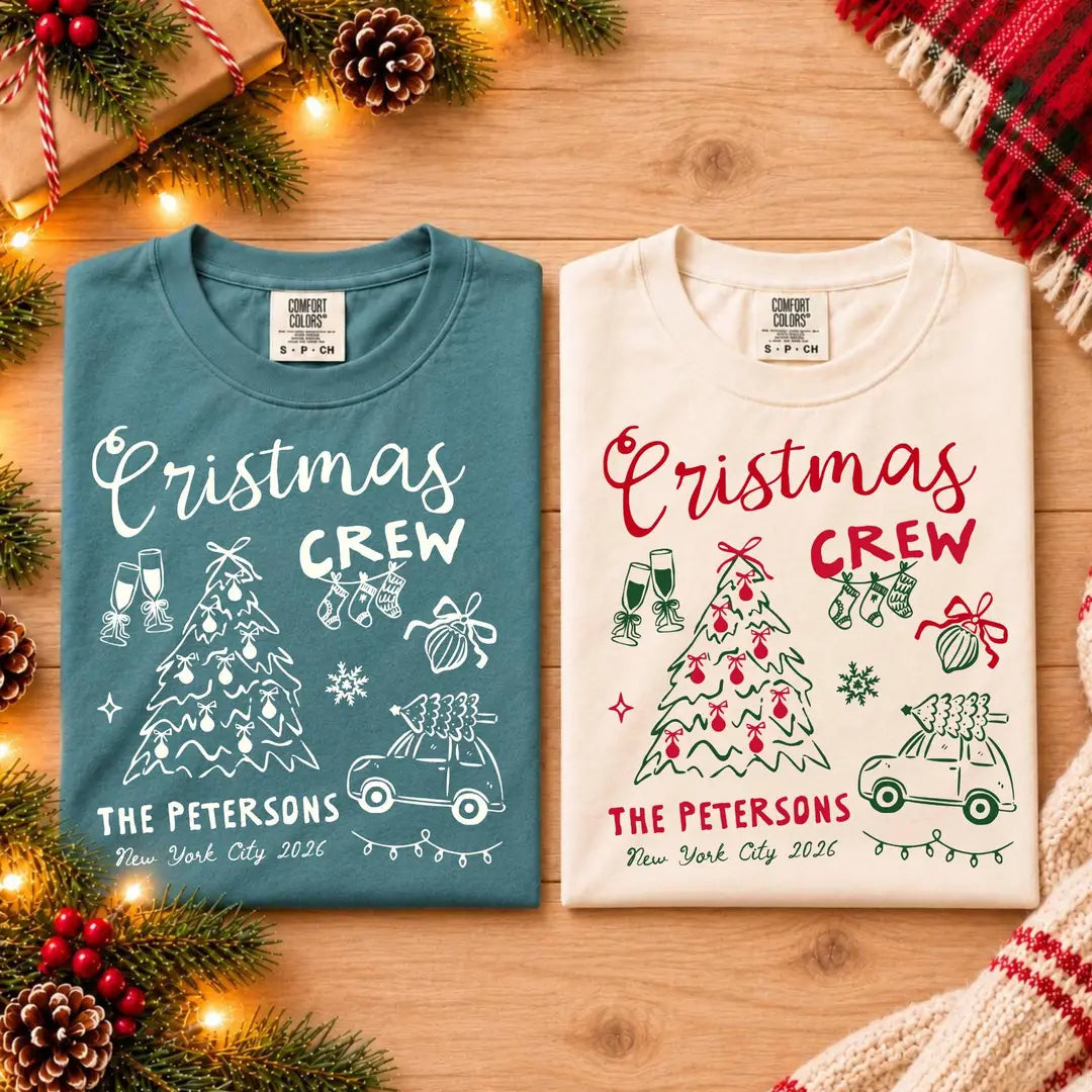 Folded Blue Spruce and Ivory Comfort Colors tees on a wood table, pine cones, crew script, bow tree, car and The Petersons