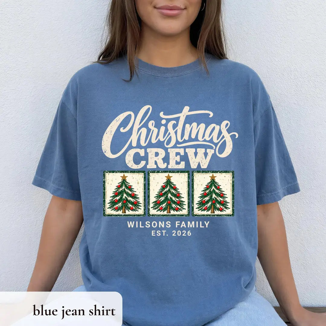 Woman in a Blue Jean Comfort Colors tee seated against a white wall, cream Christmas Crew above three trees and Est. 2026