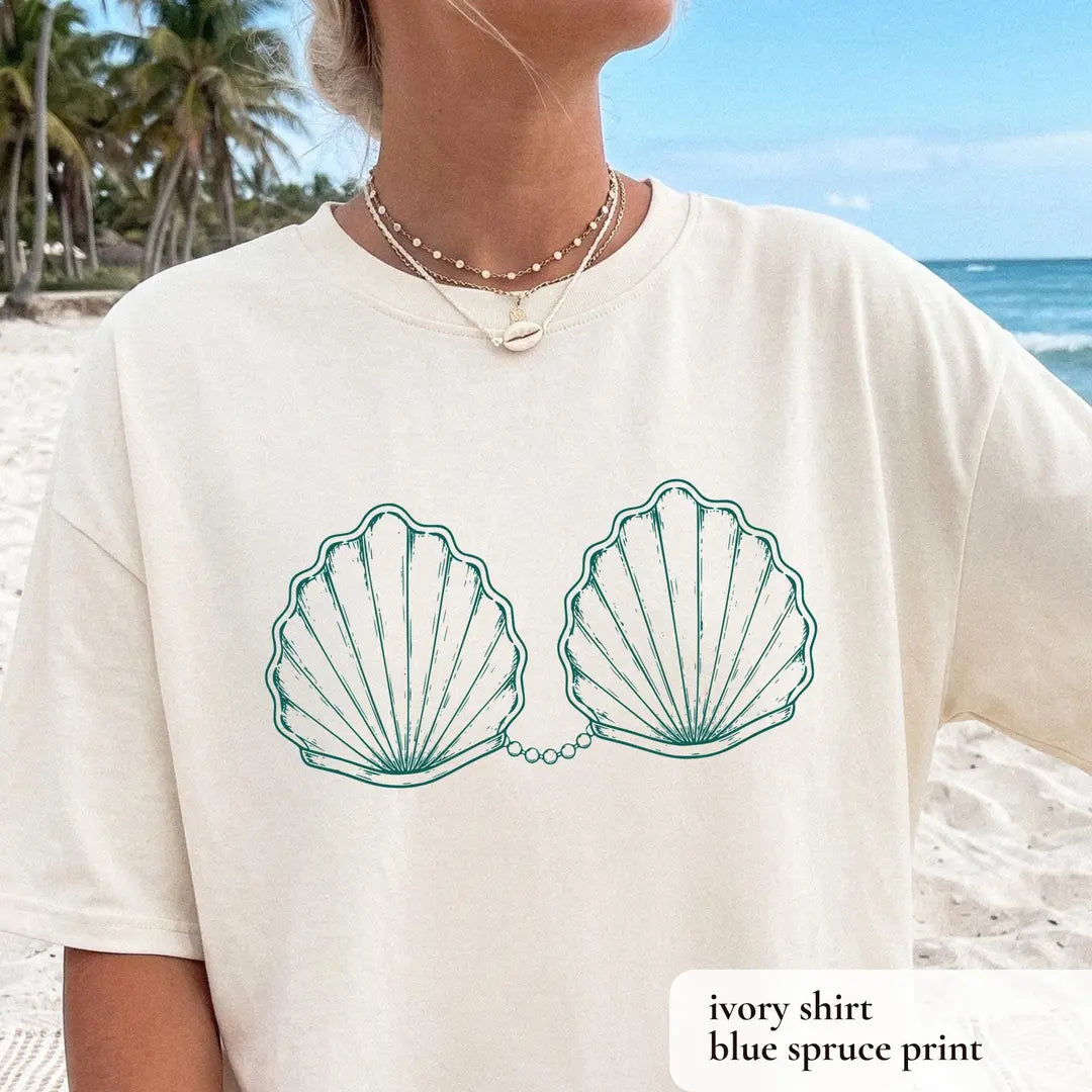 Woman in an Ivory Comfort Colors tee with palm trees and ocean behind her, two teal scallop shells linked by pearls