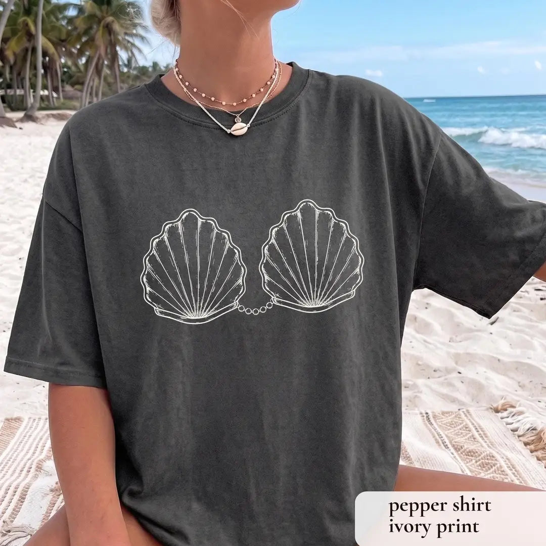 Woman in a Pepper Comfort Colors tee on a palm beach with white outlined scallop shells joined by a pearl chain