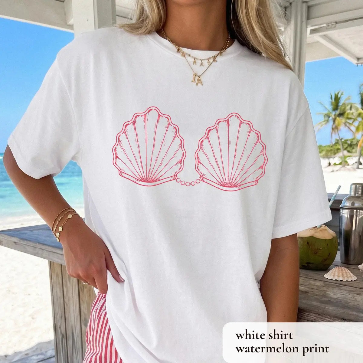 Woman in a White Comfort Colors tee at a beach bar, pink outlined scallop shells joined by a pearl chain