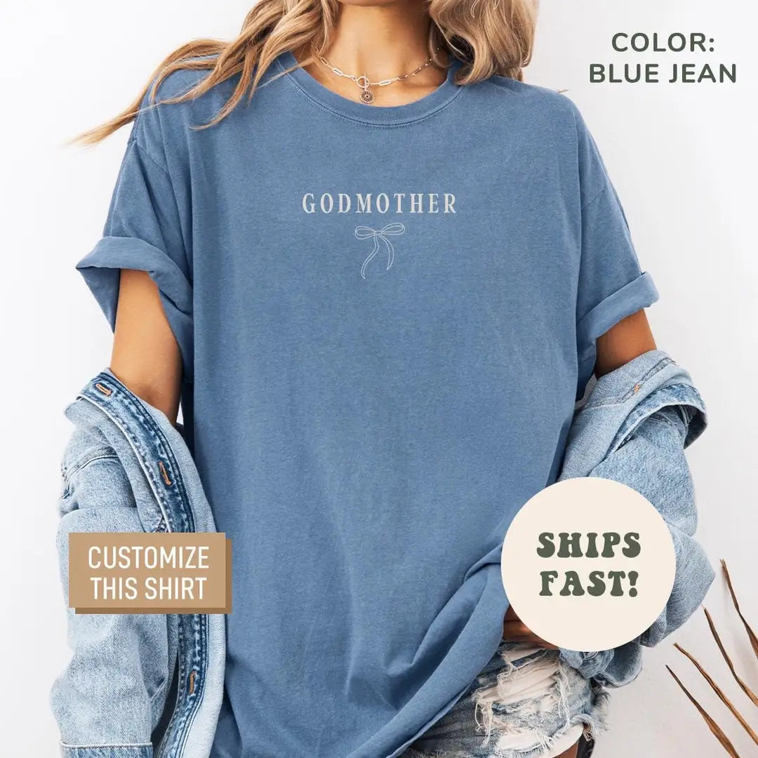 Woman in a Blue Jean Comfort Colors tee with a denim jacket off her shoulders, Godmother text over a thin bow