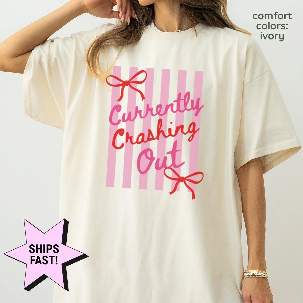 Woman in an Ivory Comfort Colors tee, hand at her hair, with red cursive currently crashing out and bows on pink stripes
