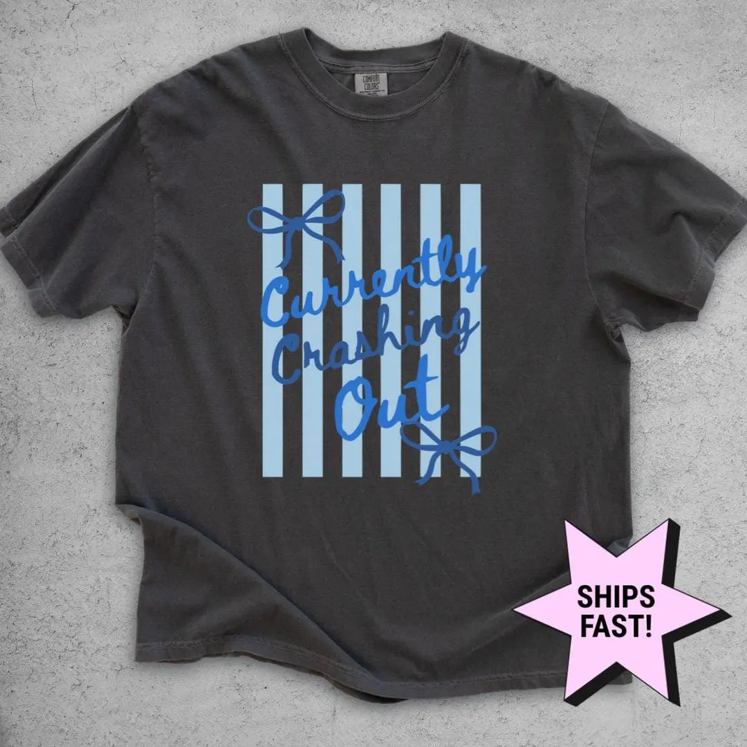 Pepper Comfort Colors tee flat on gray concrete with blue cursive currently crashing out and bows over light blue stripes