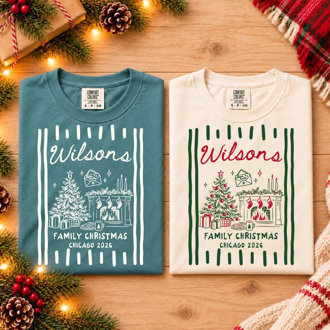 Folded Blue Spruce and Ivory Comfort Colors tees on a wood table among pine cones, Wilsons with a tree and fireplace