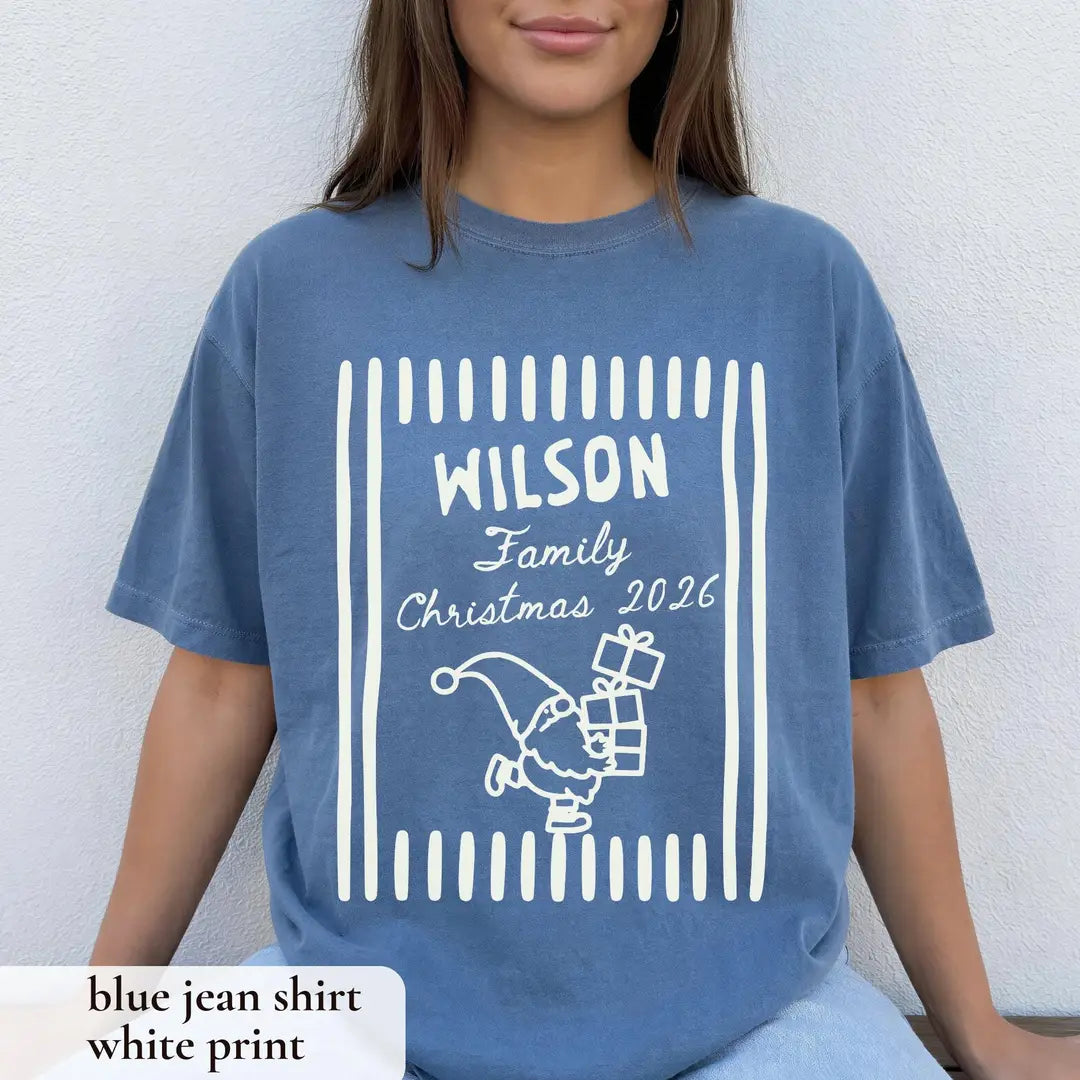 Woman in a Blue Jean Comfort Colors tee against a white wall, white Wilson Family Christmas 2026 frame, gnome with gifts