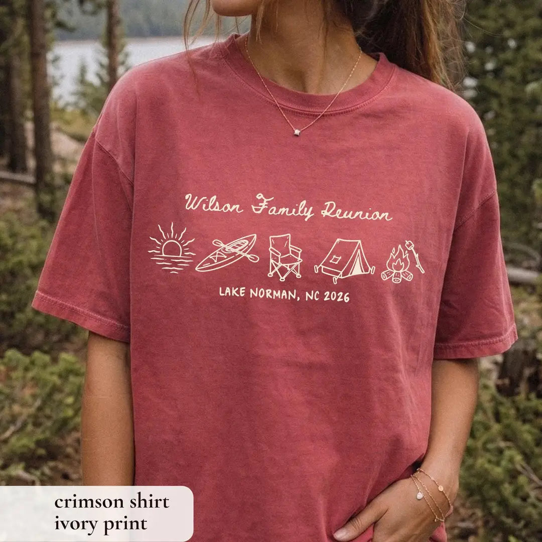 Woman in a Crimson Comfort Colors tee among pines, ivory Wilson Family Reunion script, kayak, chair, tent and campfire