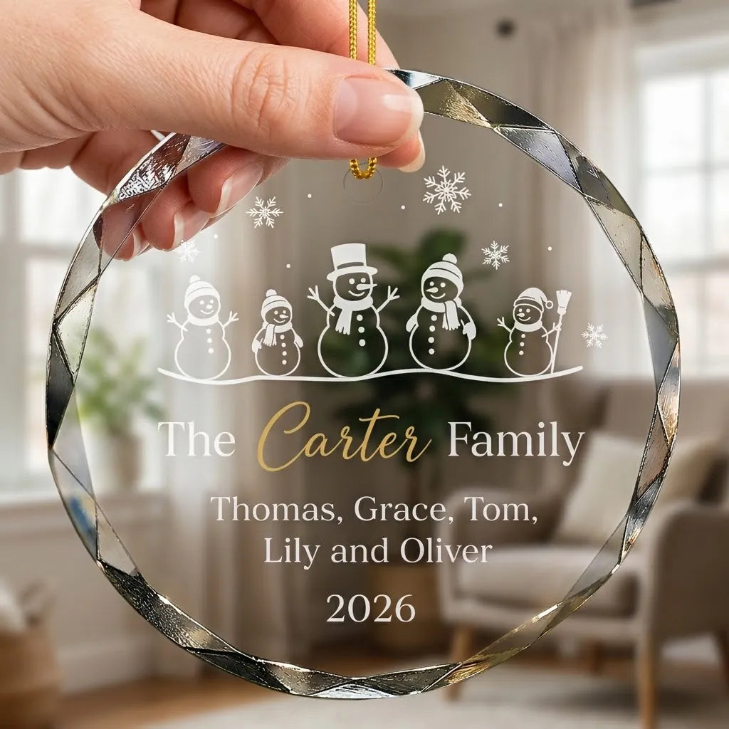 Hand holding a transparent faceted glass ornament with gold string loop showing printed five cute snowmen family line design and crisp white lettering in cozy indoor scene