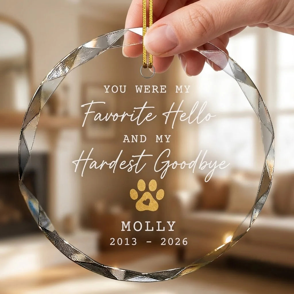 Hand holding a transparent faceted glass ornament with gold string loop showing printed gold dog cat paw print with heart design and crisp white lettering in cozy indoor scene