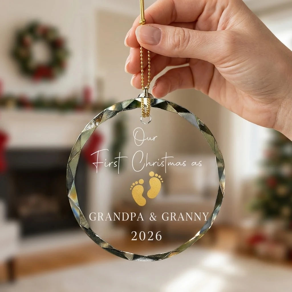 Hand holding a transparent faceted glass ornament with gold string loop showing printed gold baby footprint pair design and crisp white lettering in cozy indoor scene