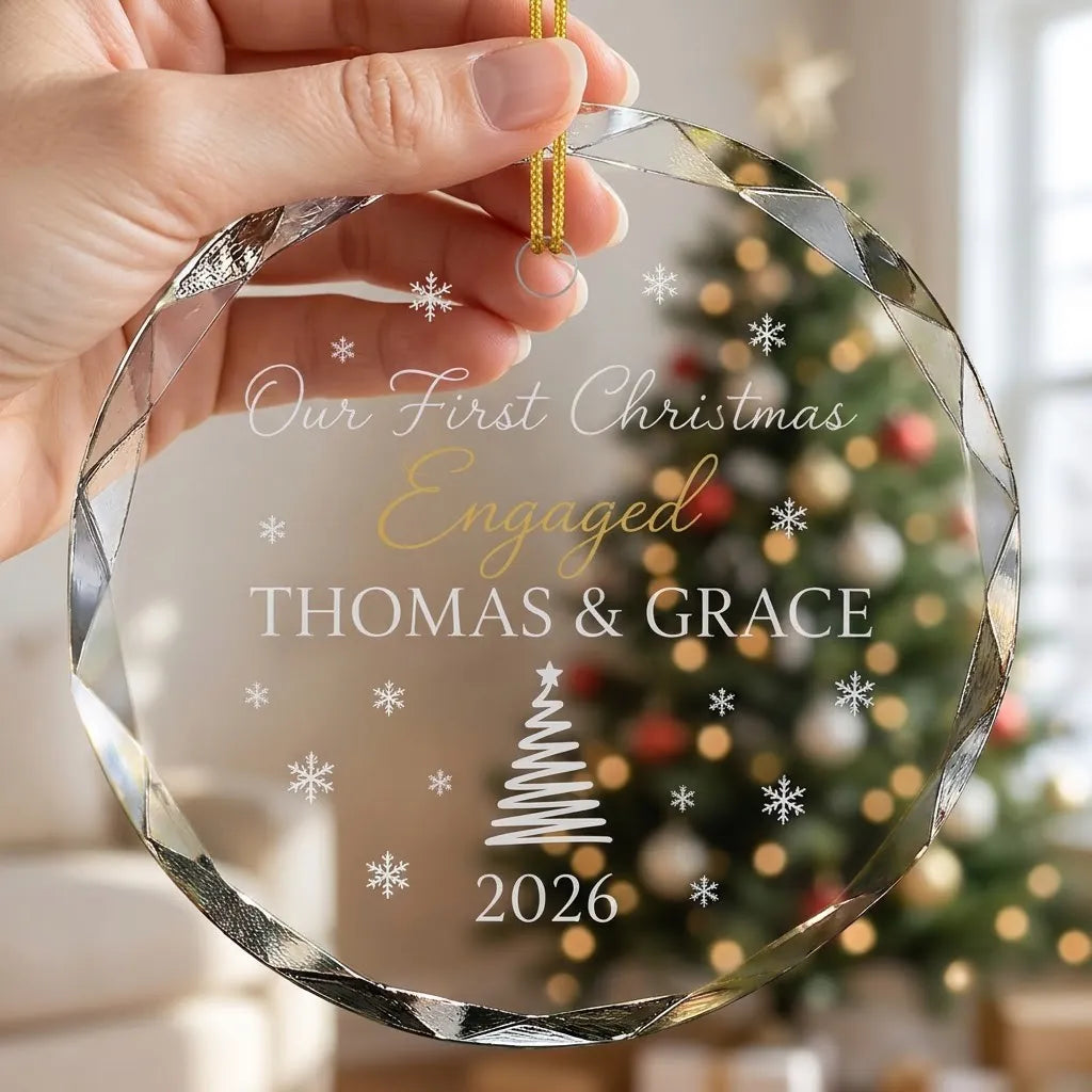 Hand holding a transparent faceted glass ornament with gold string loop showing printed pine tree and snowflakes design and crisp white lettering in cozy indoor scene