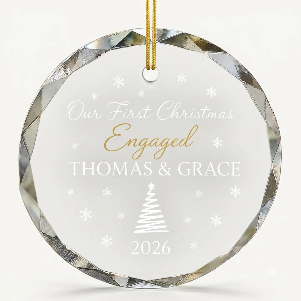 Beveled edge clear glass round ornament featuring printed pine tree and snowflakes graphics, custom text Our First Christmas Engaged and personalized details hanging on gold ribbon