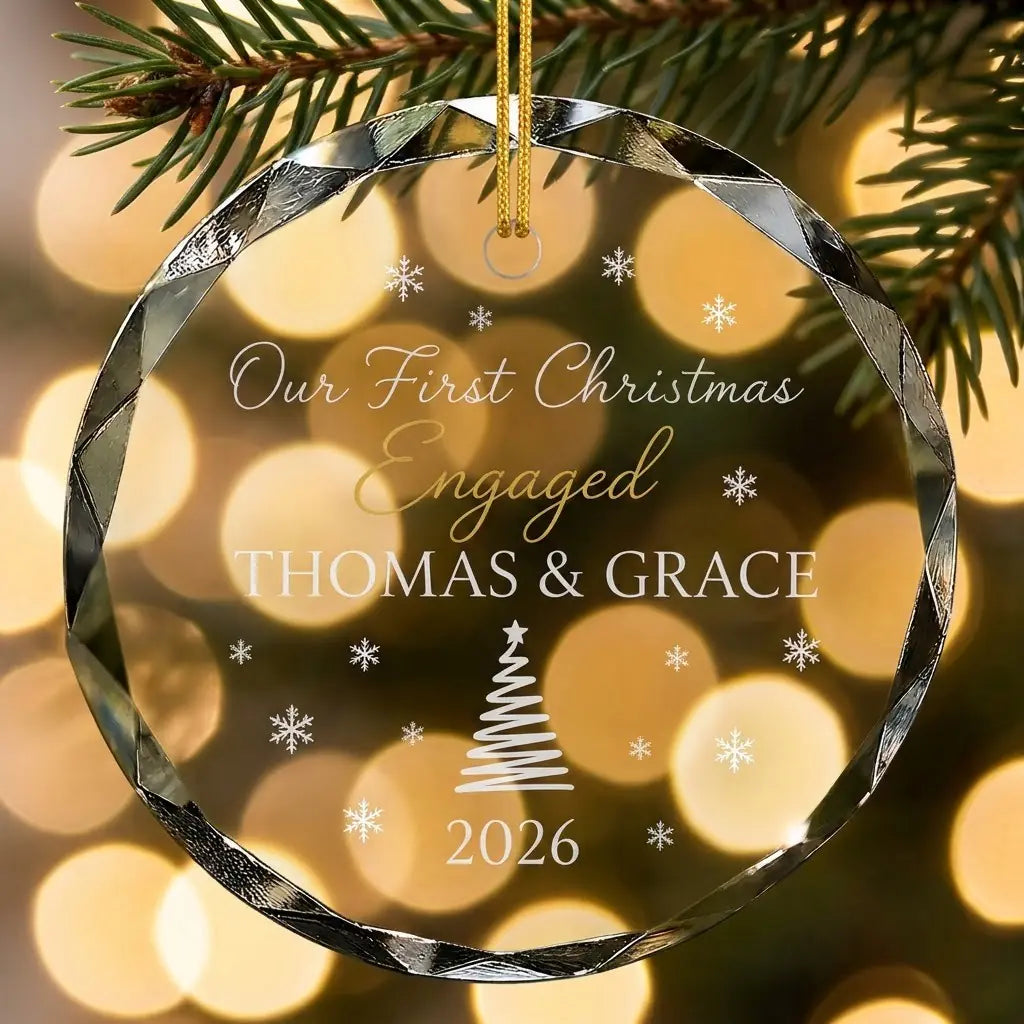 Personalized clear glass engagement ornament displayed hanging on Christmas tree branch with warm sparkling holiday lights background