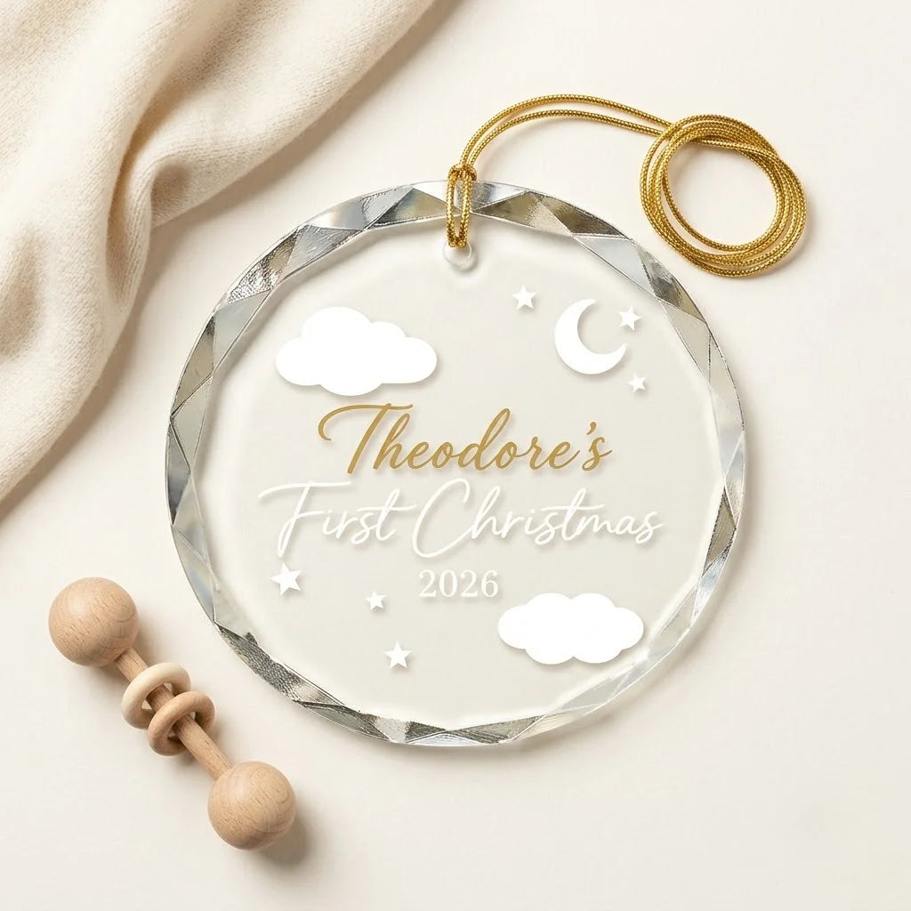 Clear glass round ornament laid flat on festive wooden surface with pine branches, pinecones and holiday decorations surrounding the baby first christmas design