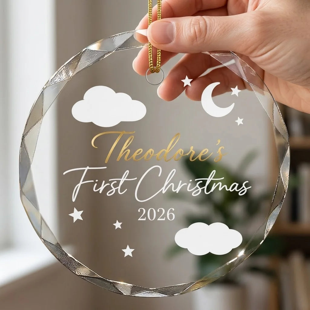 Hand holding a transparent faceted glass ornament with gold string loop showing printed crescent moon clouds and stars design and crisp white lettering in cozy indoor scene
